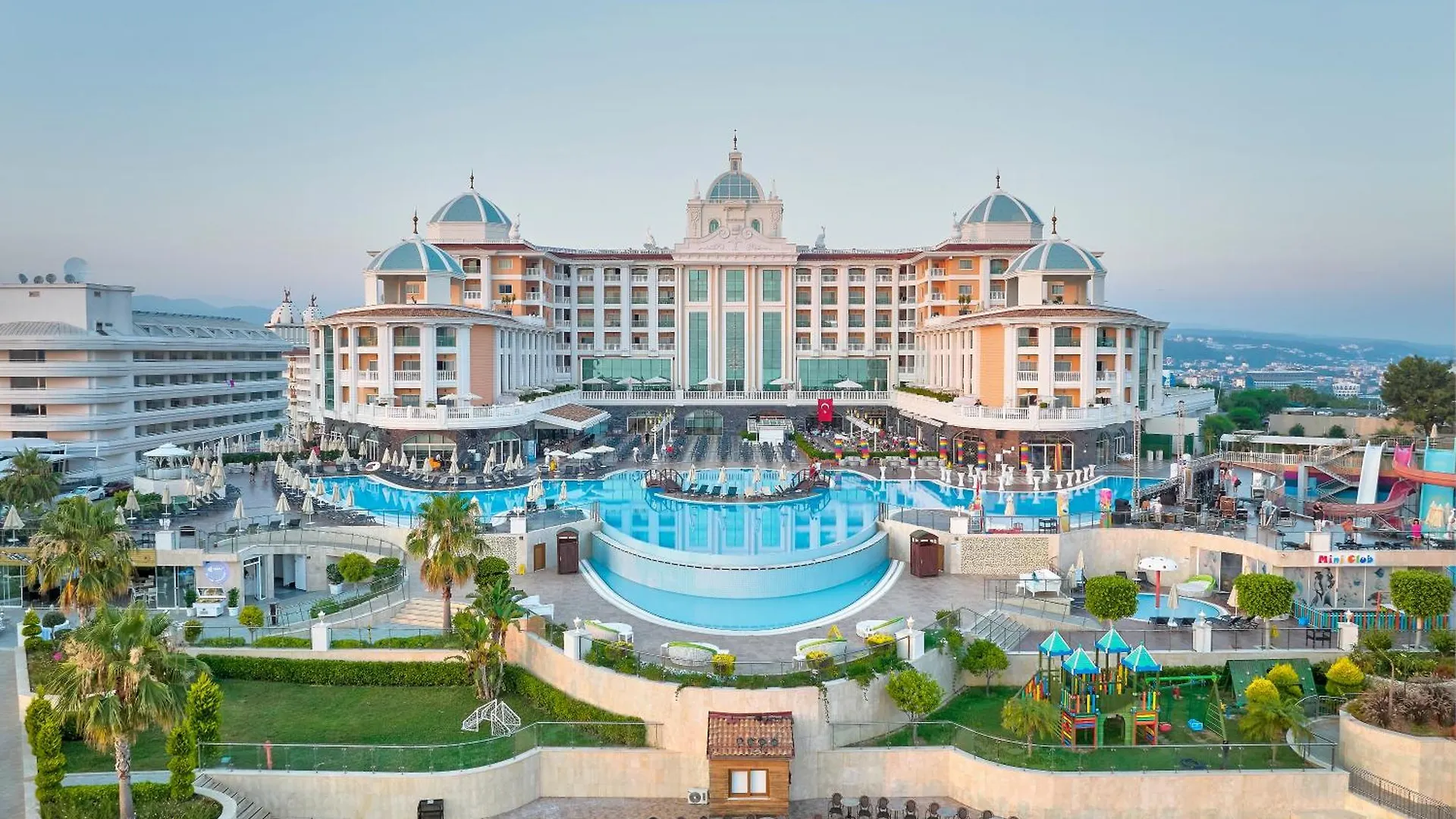 Litore Resort Hotel & Spa Okurcalar Exterior photo This is a bright, panoramic photograph showcasing a large, luxurious resort complex under a clear, light-blue sky, likely during the late morning or early afternoon.

The focal point is the grand, sprawling hotel building, which features multiple sto